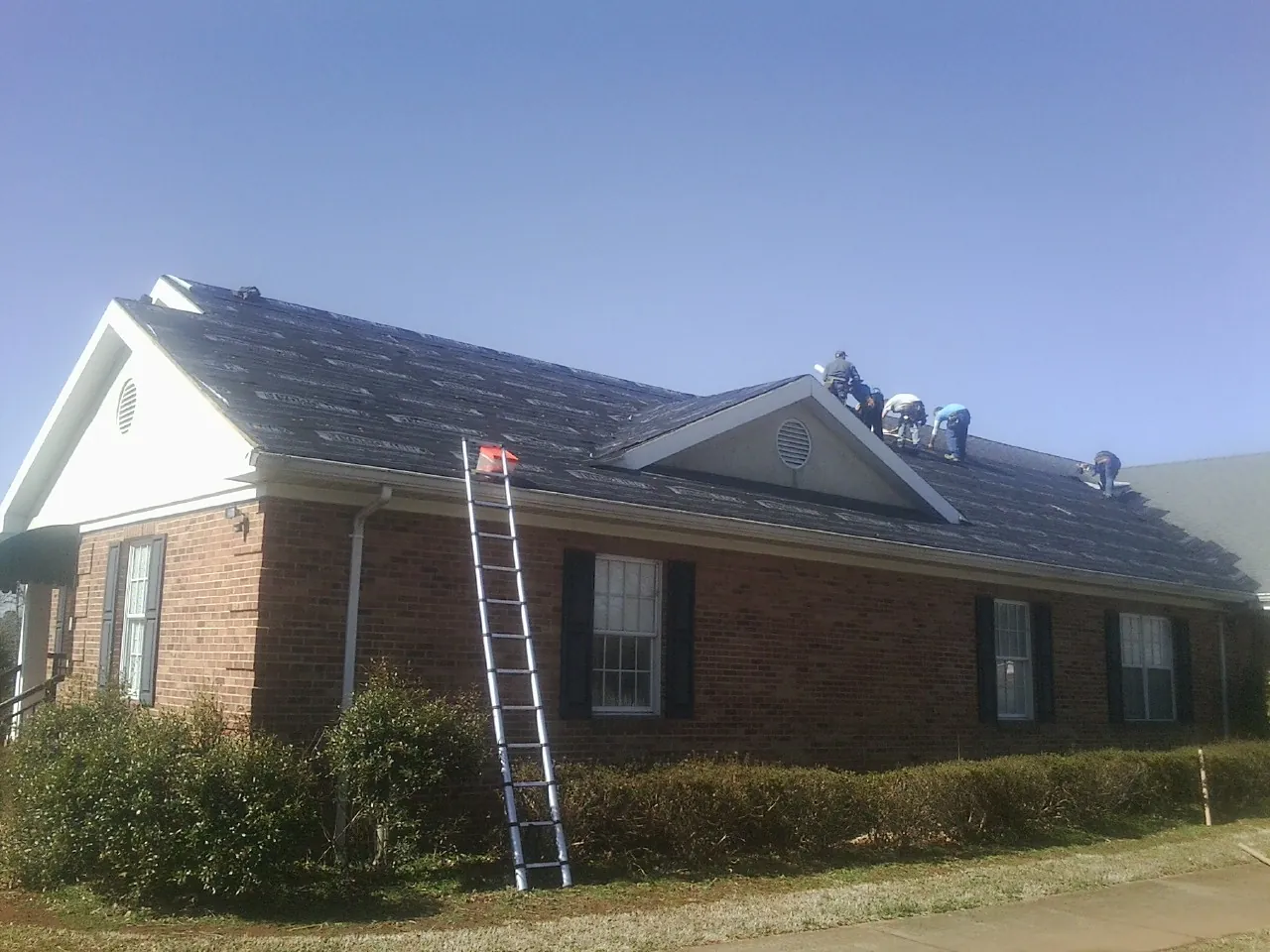 Crew tearing shingles off a brick ranch roof with a ladder set — Metal Roof Repair work by our crew in Reidville