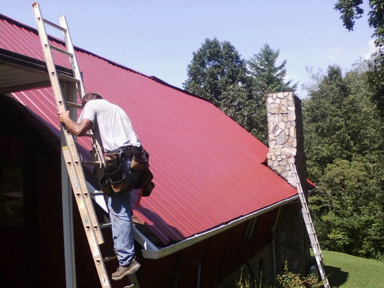Roofer on a ladder beside a red metal roof and stone chimney — Soffit & Fascia Repair by Roofing Brothers in Reidville